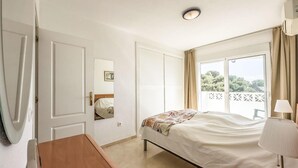 Ein Schlafzimmer mit Bett, Nachttisch, Lampe und einem Fenster mit Blick auf Grünflächen.
