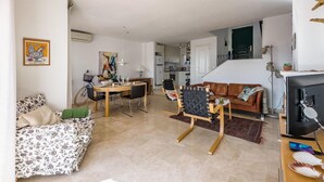 Ein Wohnzimmer mit einer Couch, einem Esstisch mit Stühlen, einer Küche im Hintergrund und einer Treppe, die in eine andere Ebene führt.
