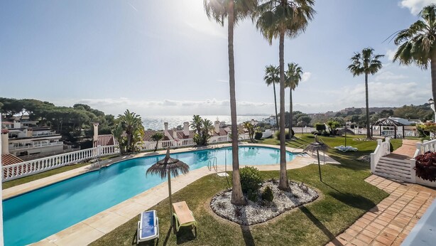 Ein Schwimmbecken mit Liegestühlen, Palmen und Blick auf das Meer und Gebäude im Hintergrund.