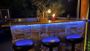 Zona de bar al aire libre con una pared de piedra, tres taburetes negros y una encimera con iluminación integrada.