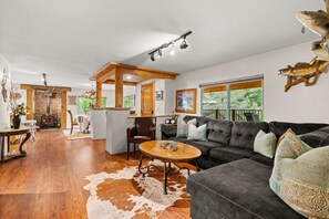 A spacious living area with a large dark-colored sofa, a wooden coffee table, and a cowhide rug. A fireplace is visible in the background, and there are large windows providing natural light.
