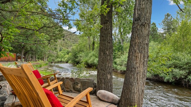 Un banco de madera con cojín rojo que da a un río, rodeado de árboles y un muro de piedra.