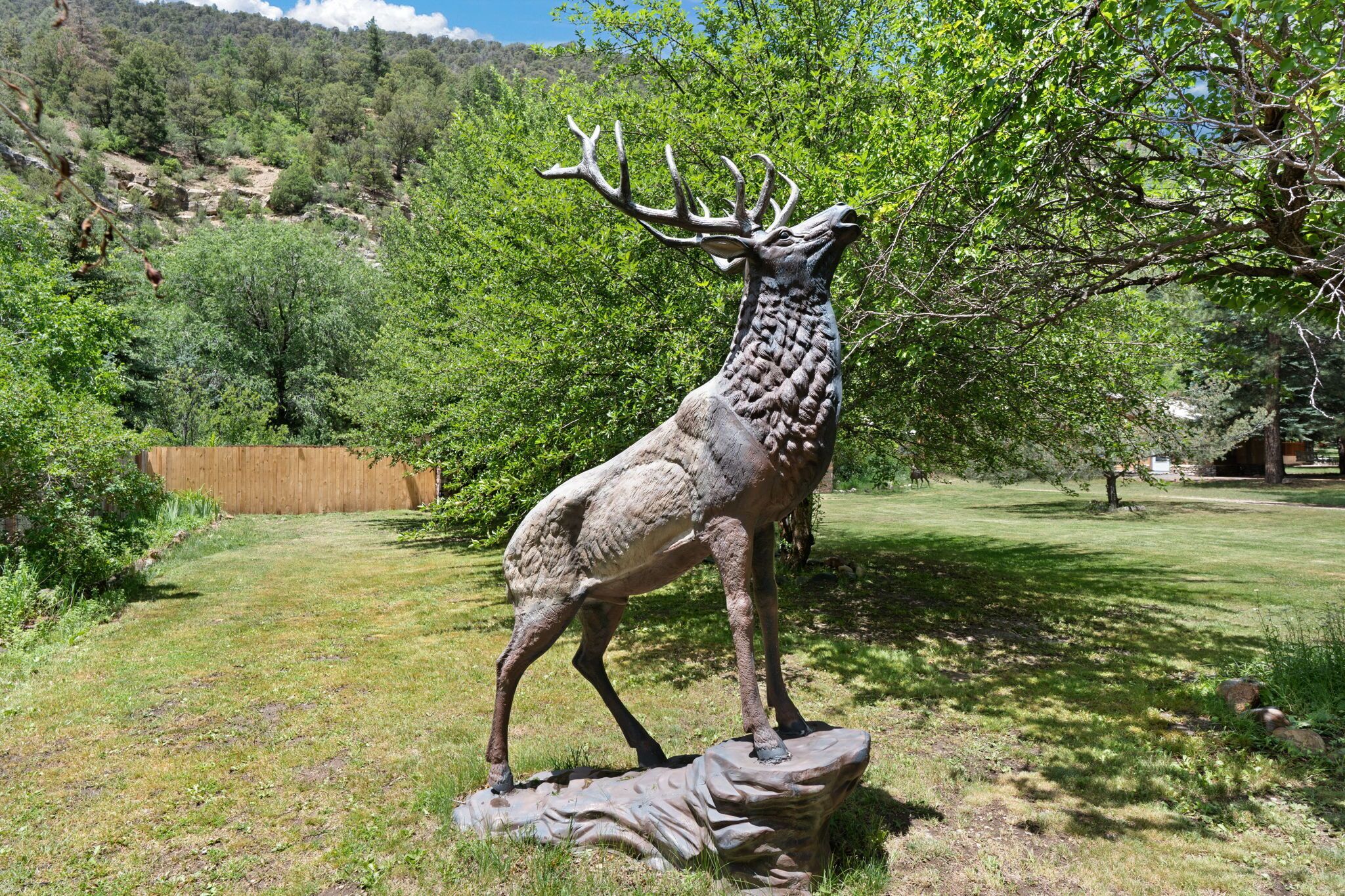 A deer statue in a grassy area with trees and a wooden fence in the background.