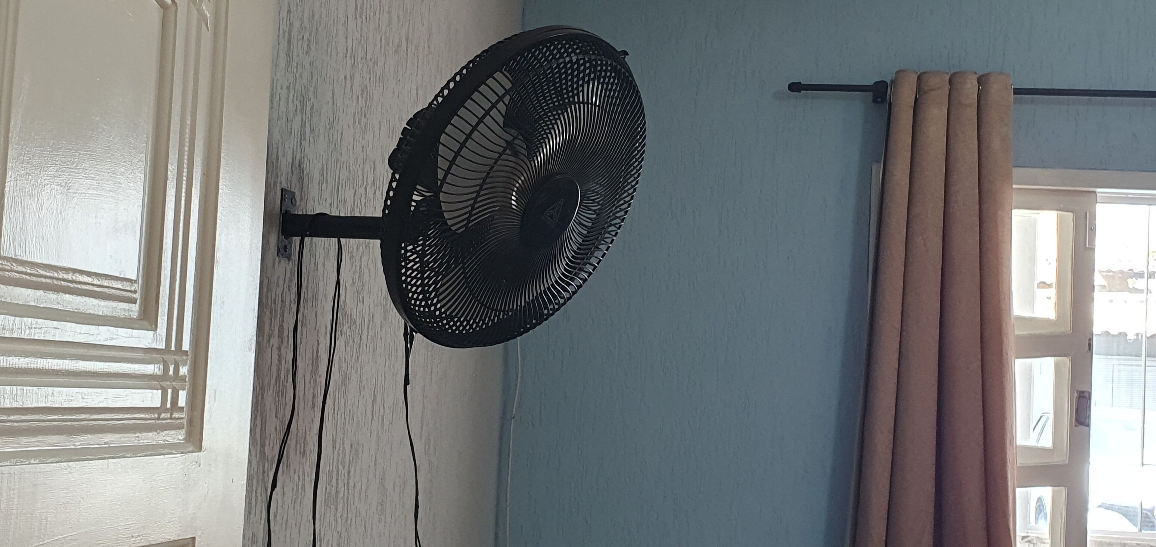 Ein Zimmer mit Standventilator, einer Tür mit dekorativer Verkleidung und einem Fenster mit Vorhängen.