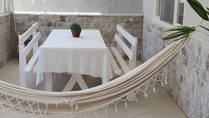 A hammock with a white table and chairs in a room with stone walls and a window. - Casa Shalom Buzios Official (Armação dos Búzios)