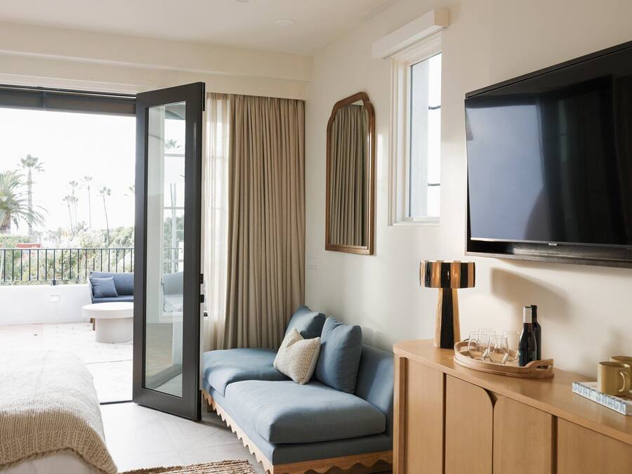 A modern hotel room with a sofa, a television, a wooden cabinet, and a balcony with a view of palm trees.
