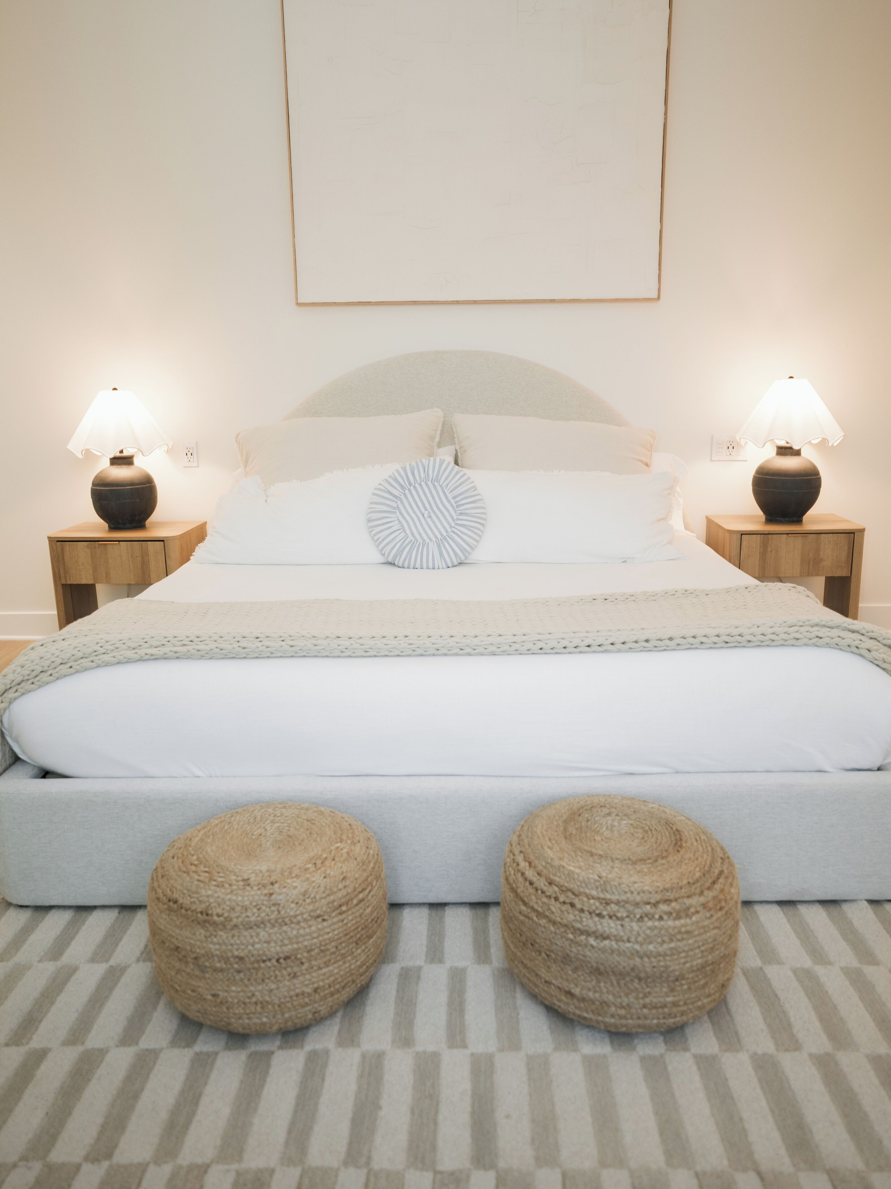 A neatly arranged bedroom with a bed, two bedside lamps, and two round ottomans on a striped carpet.