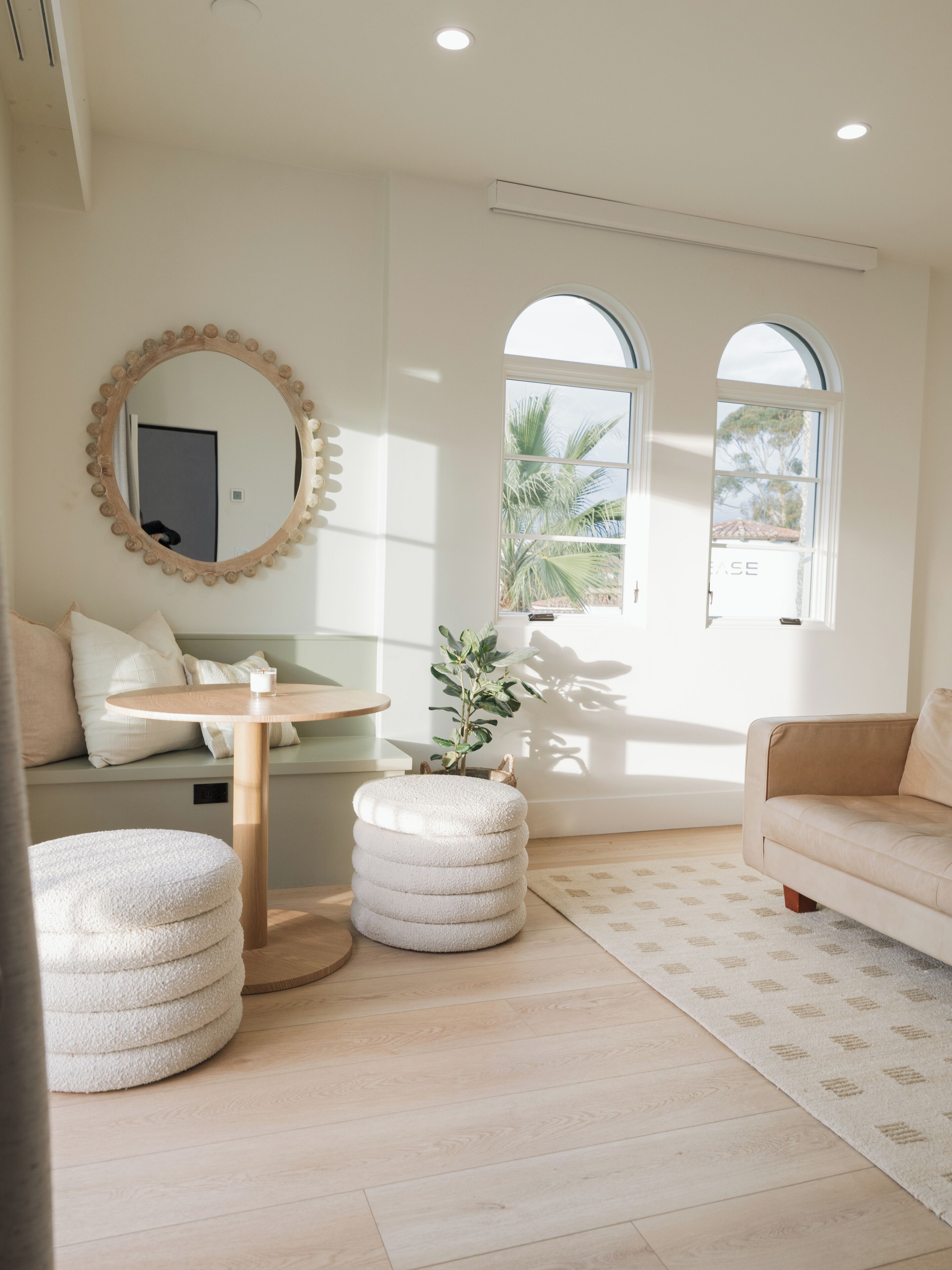 A modern living room with a round wooden table, a beige sofa, and a round ottoman. There is a large mirror on the wall and two windows with arched tops.