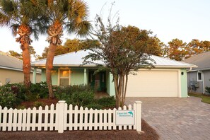 A single-story house with a garage, surrounded by a white picket fence, palm trees, and a small tree with a sign that reads "Emerald Memories."