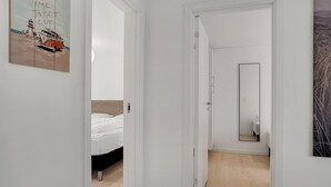 A hallway leading to a bedroom with a bed and a nightstand, a door with a mirror, and a wall with a painting.