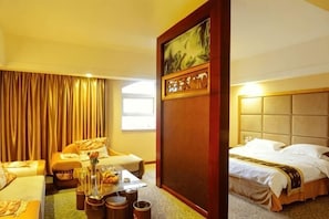 A hotel room with a bed, a sofa, a dining table with a vase and snacks, and a painting on the wall. - Garden Hotel (Jiangle)