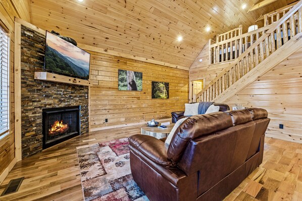 A log cabin interior with a fireplace, leather sofa, wooden walls, and a staircase. - Hot tub flat roads new build (Gatlinburg)