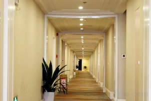 A long, narrow hotel corridor with a carpeted floor, white walls, and a ceiling with recessed lighting.