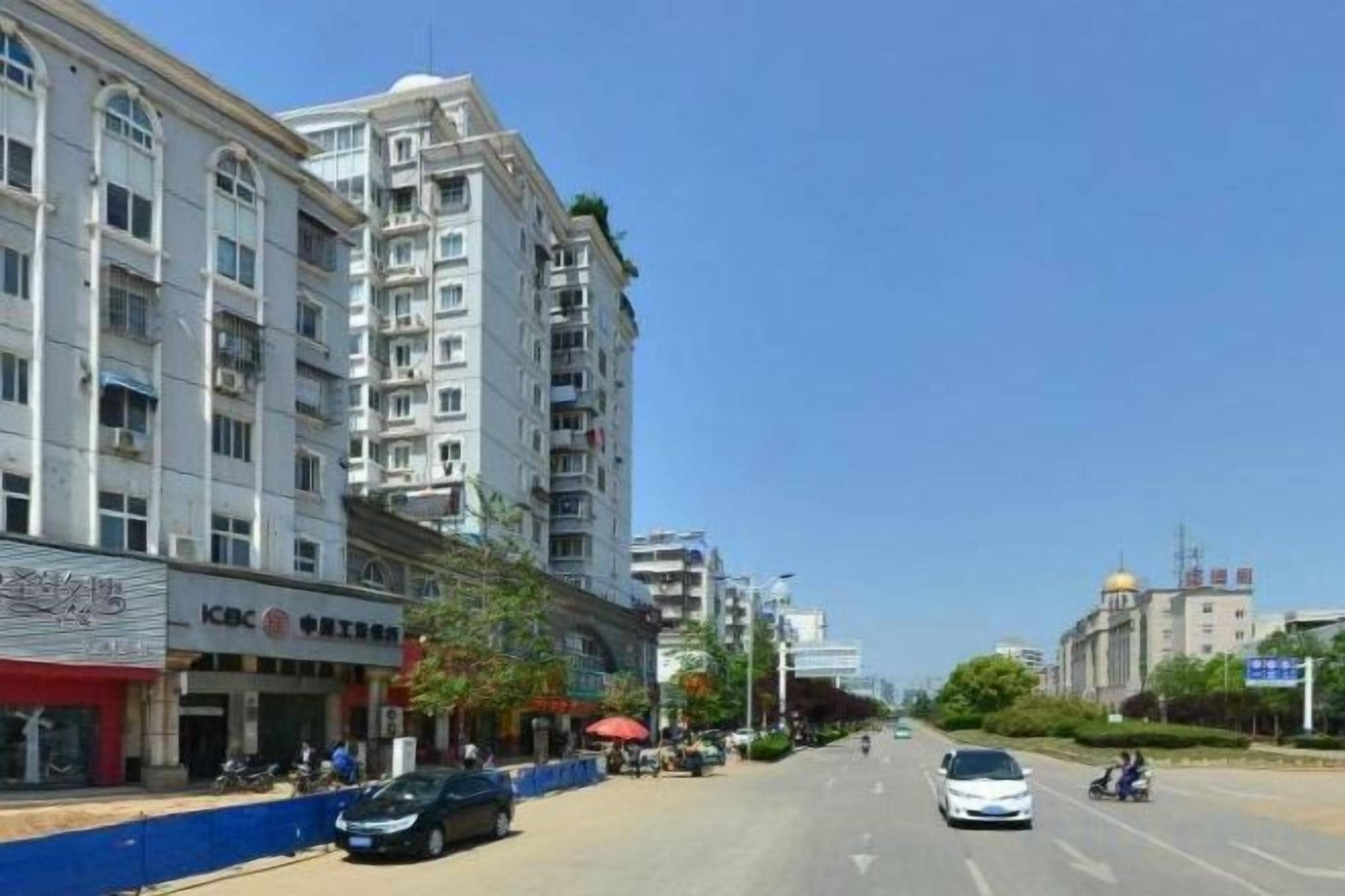 Un edifici d'alguns pisos amb locals comercials a la planta baixa, cel serena i un carrer amb vehicles.