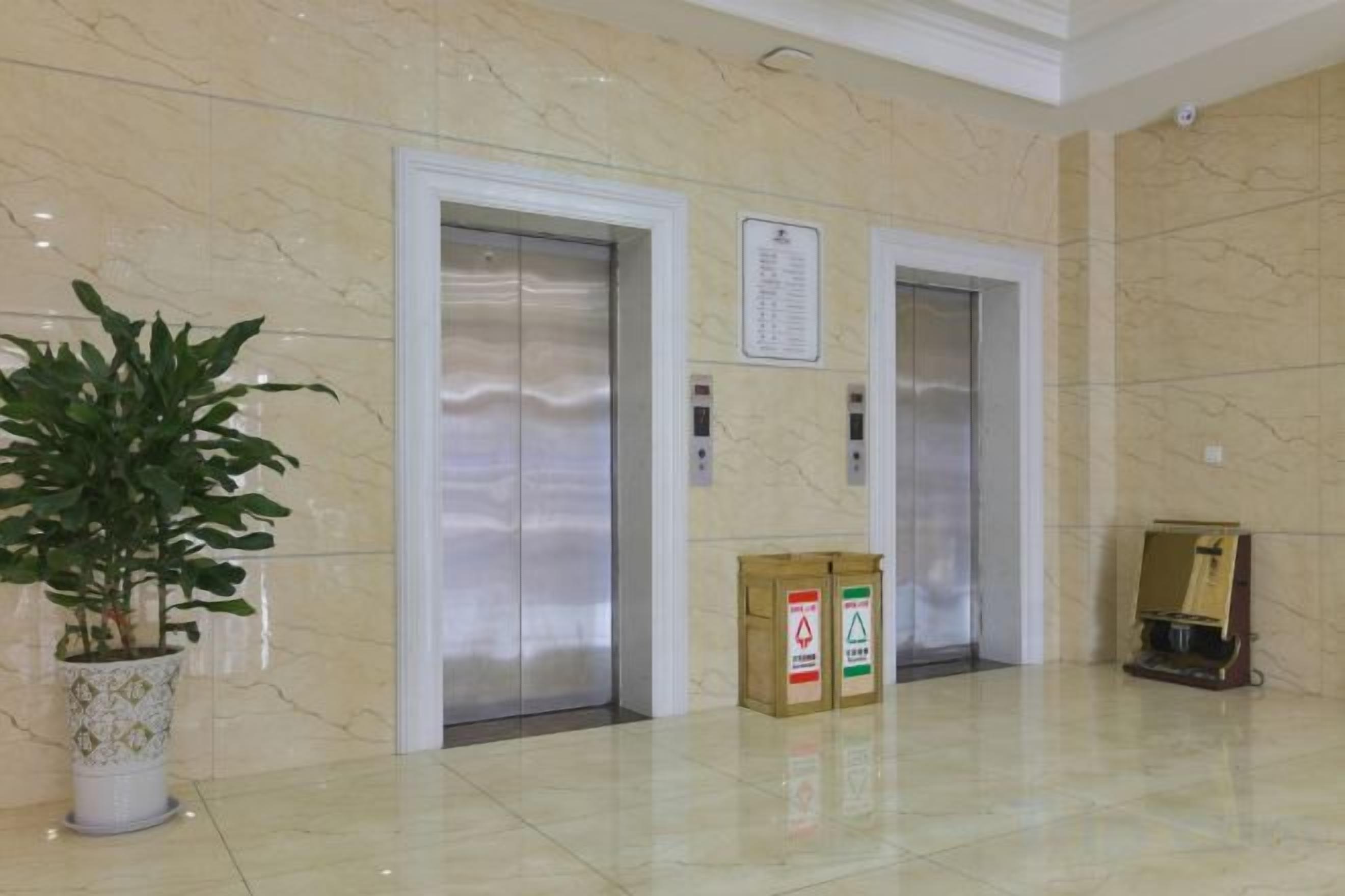 Two elevators in a lobby with a potted plant and boxes on the floor.