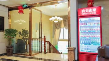 Un vestíbulo de hotel con entrada por puerta de cristal, una máquina expendedora de bebidas roja y un candelabro.