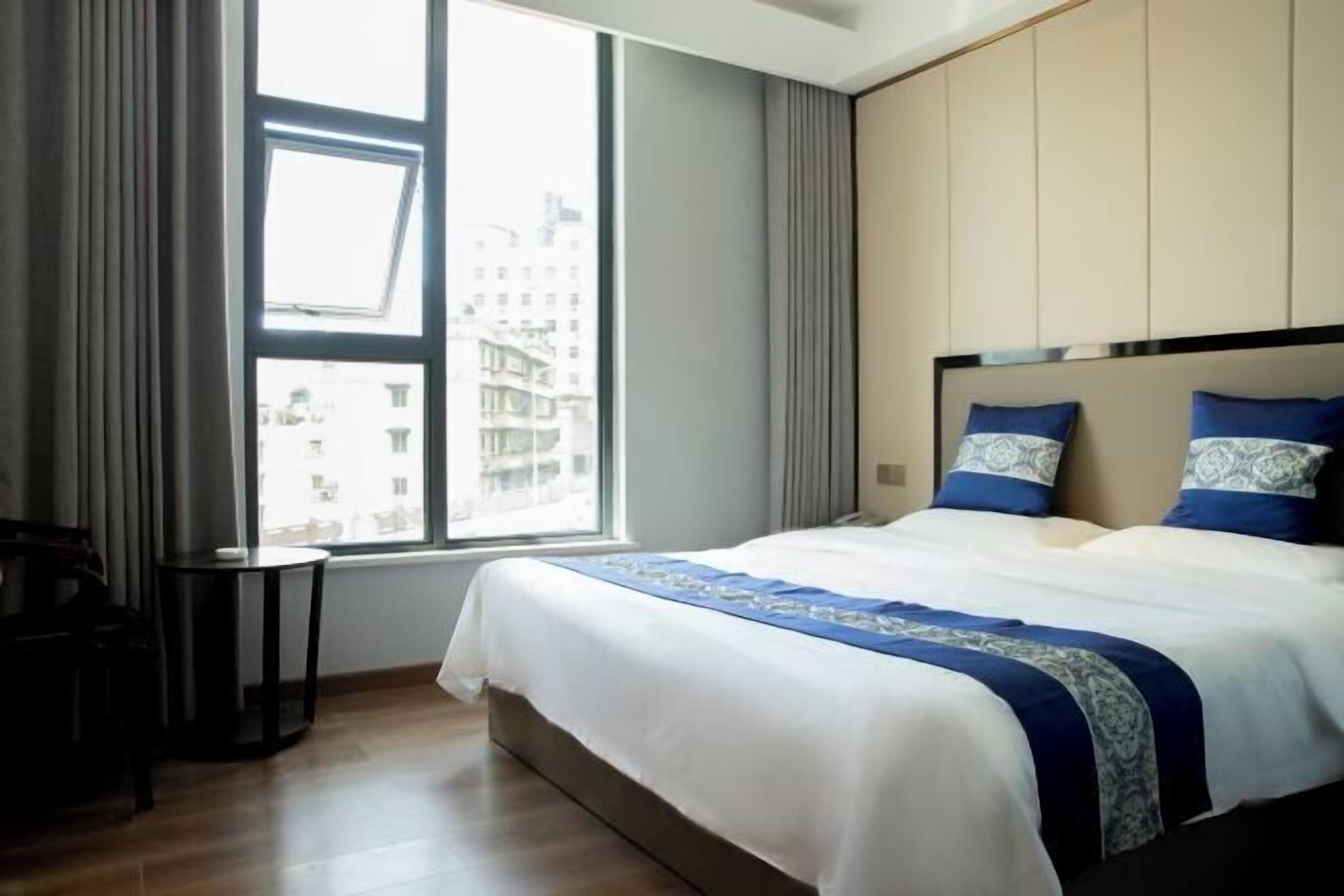 Une chambre d’hôtel avec une grande fenêtre, un lit avec une literie bleue et blanche, une petite table de chevet et un plancher en bois.
