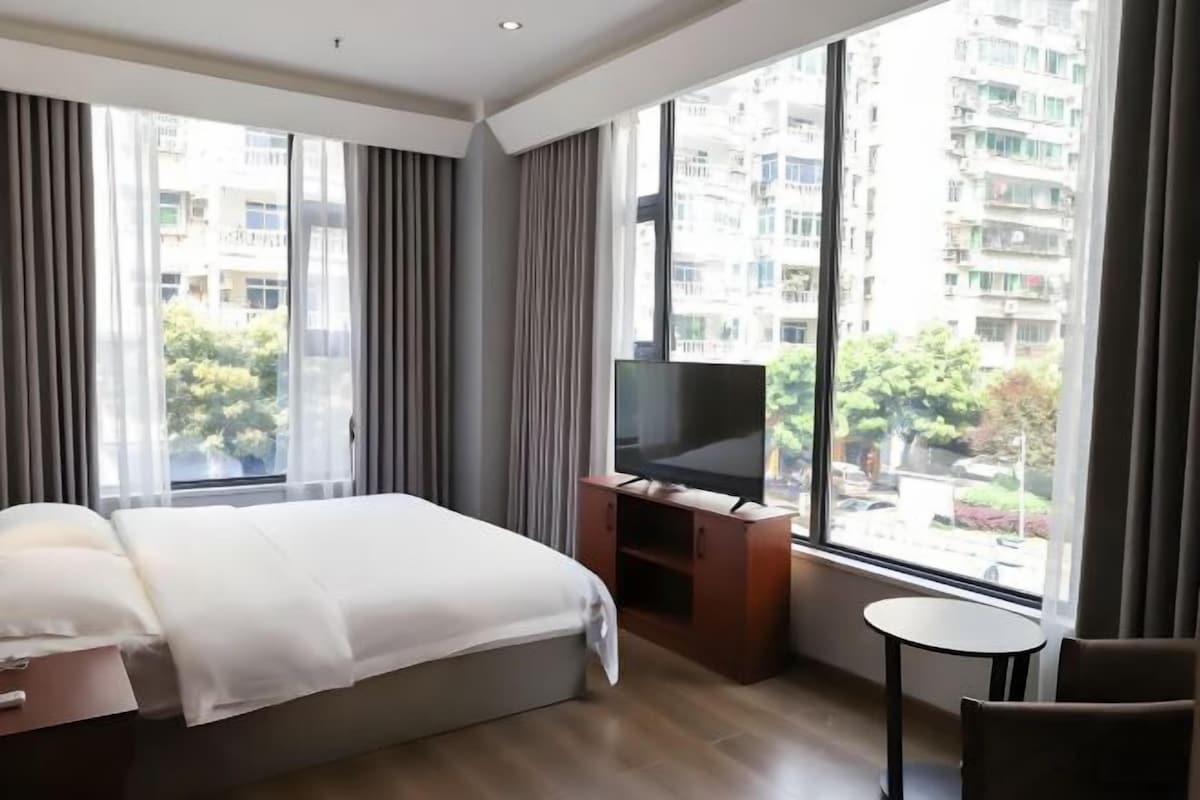 A hotel room with a bed, a TV on a wooden stand, a small round table, and a large window with curtains.