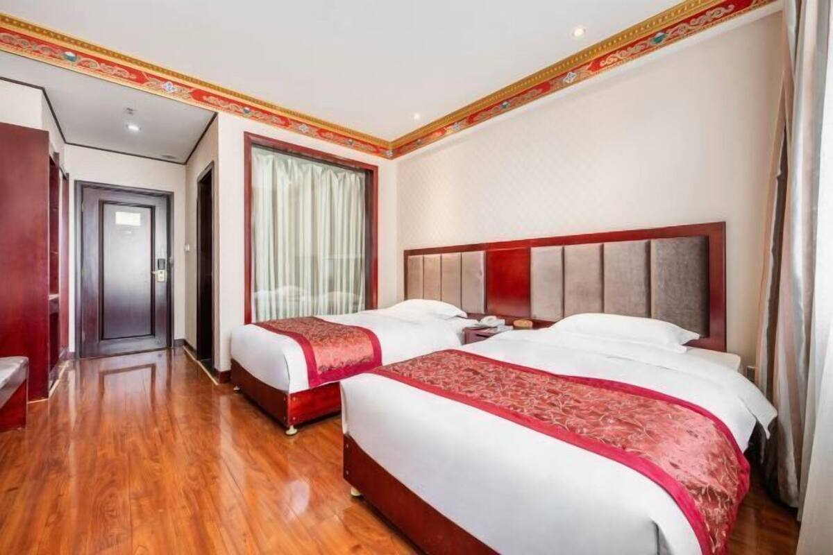Habitación de hotel con dos camas, piso de madera, colcha roja y blanca, ventana grande con cortinas y puerta que da a otra habitación.