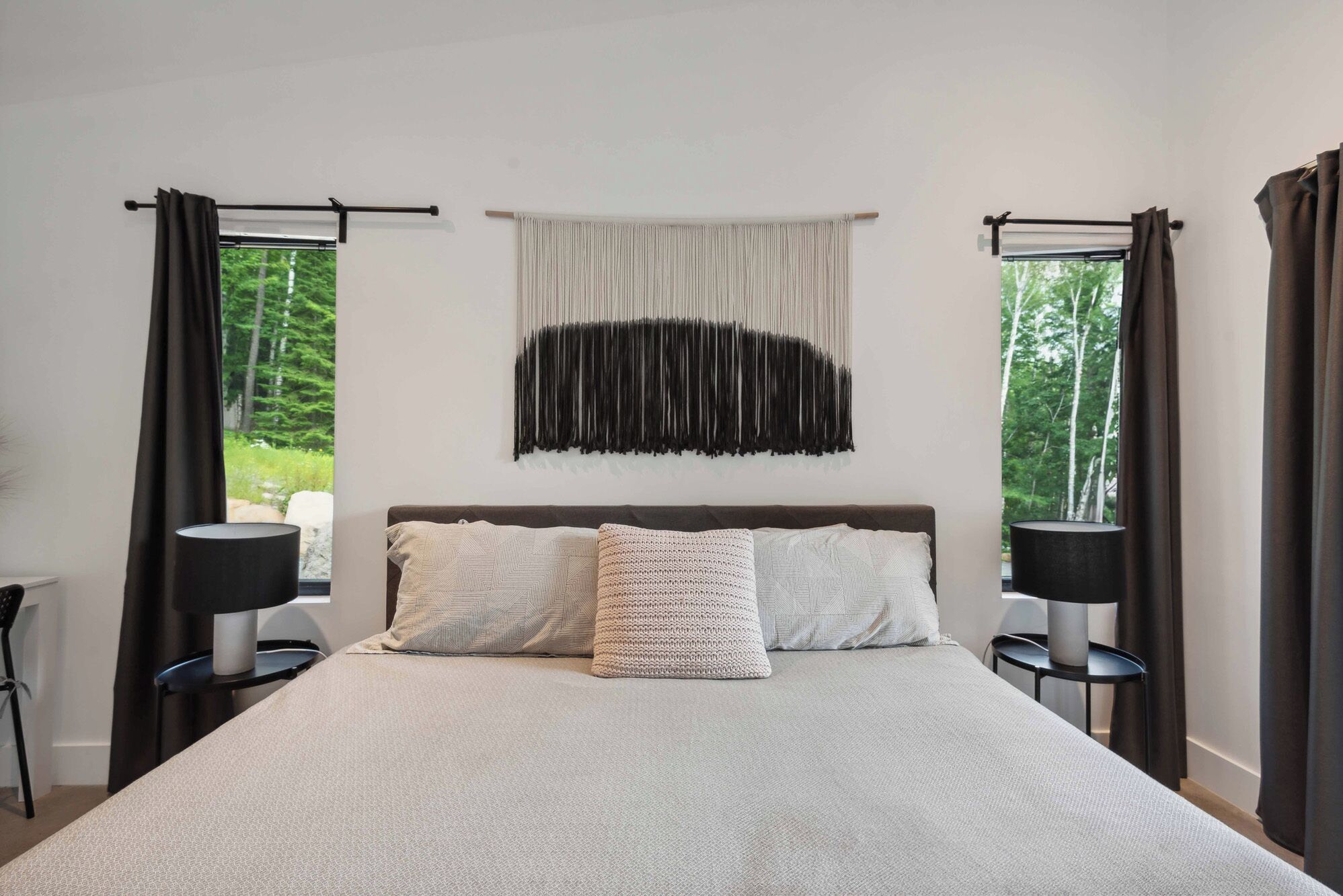 Une chambre avec un grand lit, deux tables de chevet, une tapisserie murale et une vue sur la verdure par les fenêtres.
