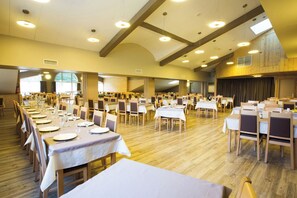 Un amplio salón de banquetes con numerosas mesas y sillas, techos altos y luz natural que entra por las ventanas.