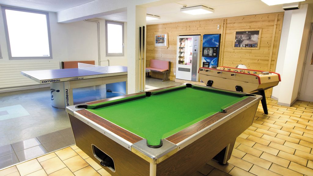 A pool table with a green felt surface, a foosball table, and a ping pong table in a room with wooden paneling and tiled flooring.