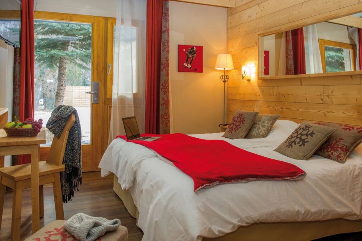 A cozy bedroom with a bed, a desk, and a chair. There is a window with curtains, a lamp, and a painting on the wall.