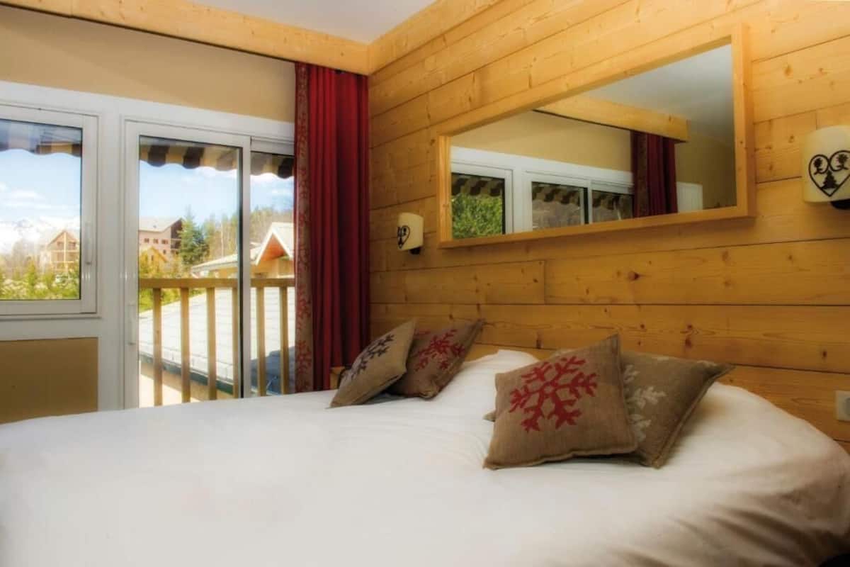 A bedroom with a bed, wooden walls, a large mirror, and a view of buildings through a sliding door.