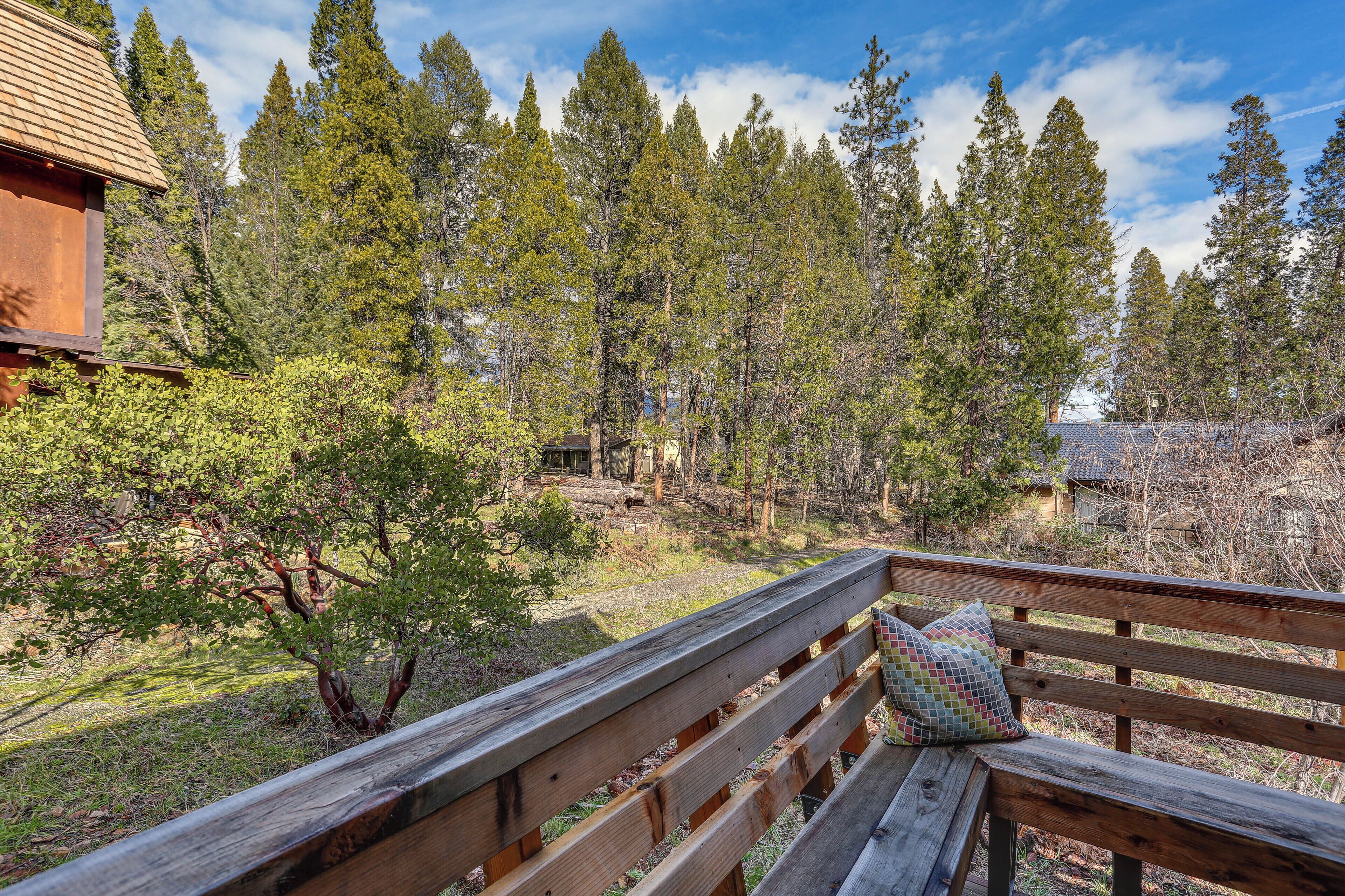 A wooden deck with a bench and a cushion, overlooking a forested area with tall trees and a cabin in the background.