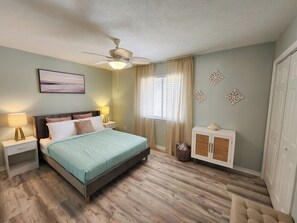 Une chambre à coucher comprenant un lit, des tables de chevet, un ventilateur de plafond, une fenêtre avec des rideaux, une armoire et une œuvre d’art accrochée au mur.