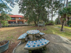 A mosaic-tiled outdoor seating area with a central table and benches, surrounded by trees and a building in the background.