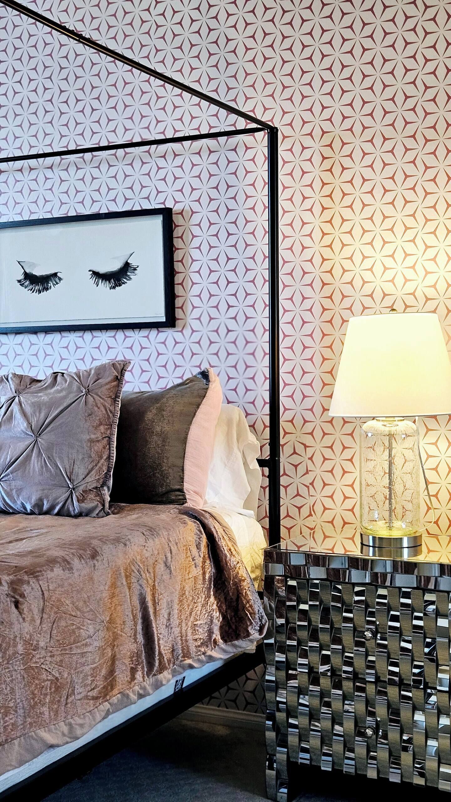 A bedroom with a geometric patterned wallpaper, a bed with multiple pillows, a framed picture above the bed, and a lamp on a side table.