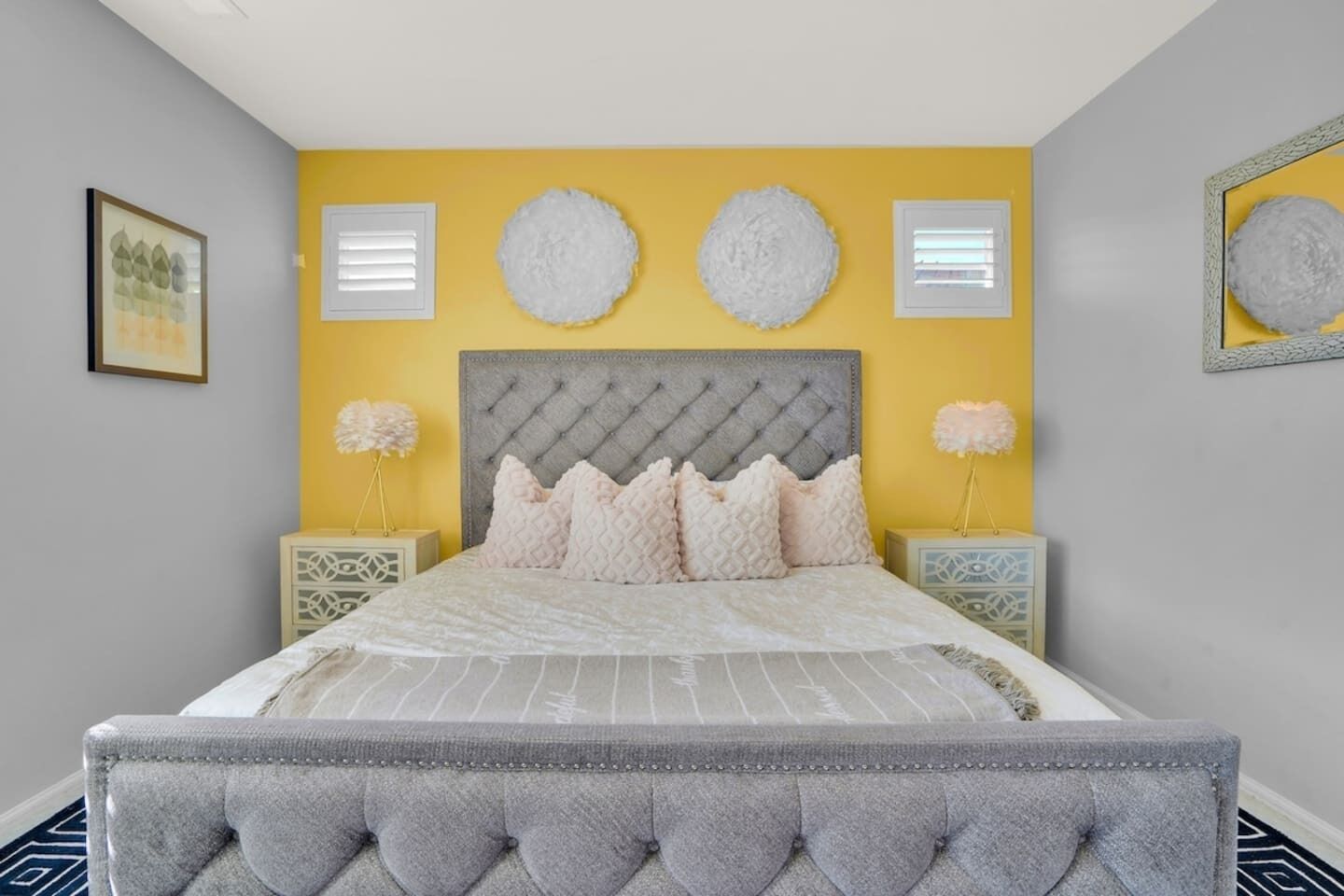 Un dormitorio con cabecera capitonada, cama con colcha a rayas blancas y grises, dos mesitas de noche con lámparas decorativas y un cuadro enmarcado en la pared.