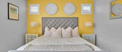 Un dormitorio con cabecera capitonada, cama con colcha a rayas blancas y grises, dos mesitas de noche con lámparas decorativas y un cuadro enmarcado en la pared.