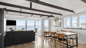 Ein moderner Wohnraum mit großem Sofa, Esstisch und Stühlen, einem an der Wand befestigten Fernseher und Meerblick durch große Fenster.