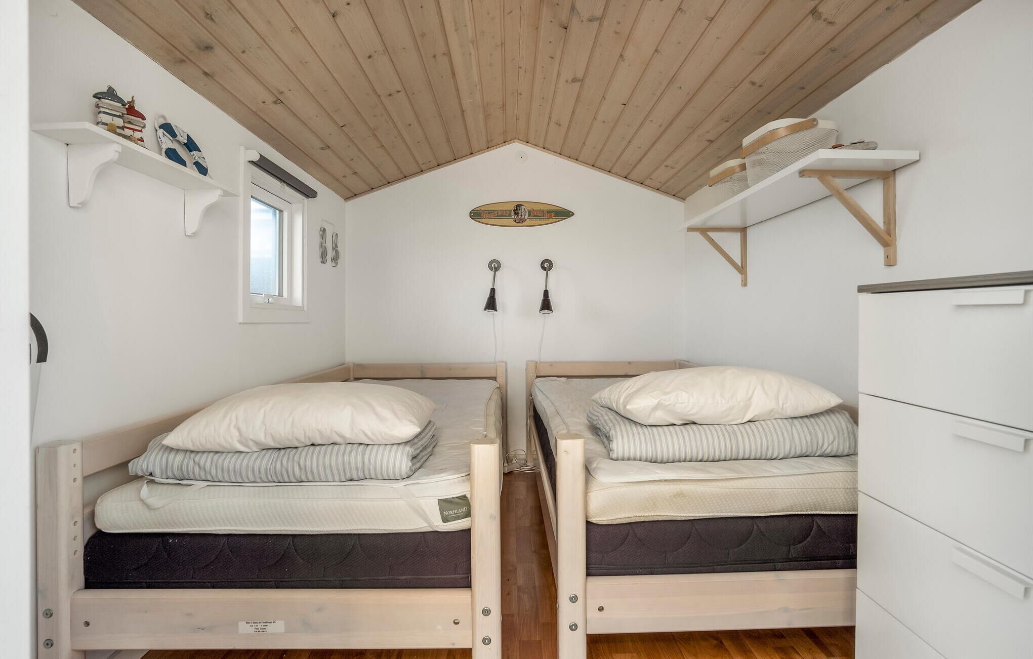 A compact bedroom with two single beds, wooden ceiling beams, white walls, and built-in storage shelves.
