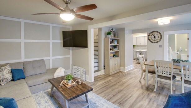 A living room with a sofa, coffee table, and a dining area with a table and chairs. There is a ceiling fan, a flat-screen TV mounted on the wall, and a staircase leading to the kitchen. - Coastal Comfort & Golfers' Paradise in Sheboygan! (Sheboygan)