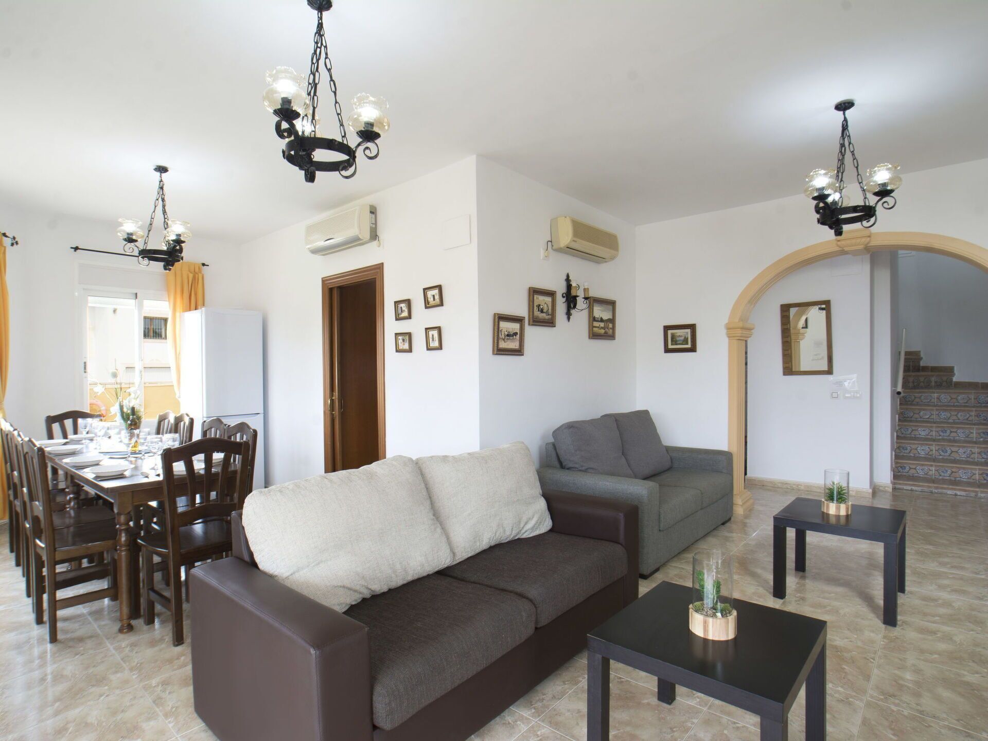 A living room with a sofa, a dining table set for four, and two chairs. There are two air conditioning units on the wall, a chandelier, and a staircase leading to another level.