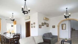 A living room with a sofa, a dining table set for four, and two chairs. There are two air conditioning units on the wall, a chandelier, and a staircase leading to another level. - Wonderful private villa for 10 people with private pool, A/C, WIFI, TV, terrace and pets allowed (Benissa)