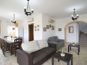 A living room with a sofa, a dining table set for four, and two chairs. There are two air conditioning units on the wall, a chandelier, and a staircase leading to another level. - Wonderful private villa for 10 guests with A/C, private pool, WIFI, TV, terrace and pets allowed (Benissa)