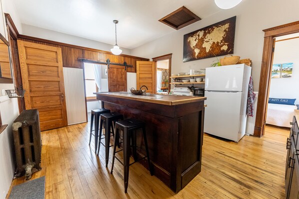 Una cocina con piso de madera, una isla central con taburetes, un refrigerador blanco y una lámpara colgante.