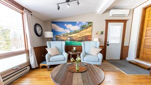 A living room with two light blue armchairs, a wooden coffee table, a large painting of a coastal landscape, a clock on the wall, and a door with a wooden frame. - Our Place Inn Munising; Munising | 5 Min to Pictured Rocks | 3BR (Munising)