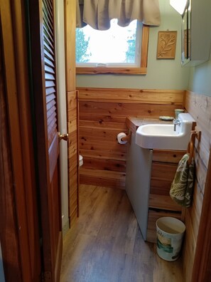 Un baño pequeño con paredes de madera, una ventana con cortina, un lavamanos y un inodoro.