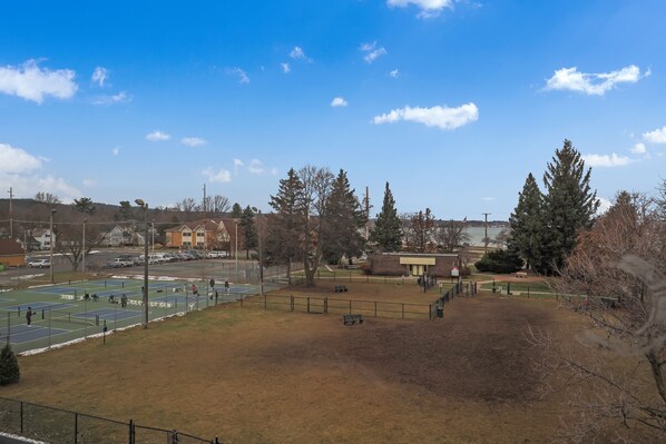 A tennis court with a fence, trees, and a building in the background.