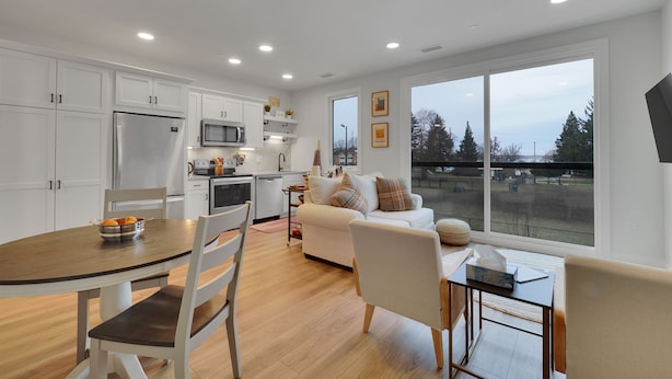 A modern kitchen with white cabinetry, a stainless steel refrigerator, and a microwave. There is a dining table with chairs and a bowl of oranges. A white sofa is positioned near a large window with a view of trees and buildings. - The Bish: Traverse City Fall Getaway | Wine Country & Downtown | Sleeps 2 (Traverse City)