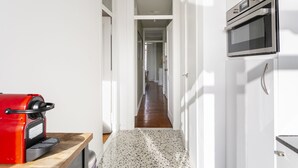 Un couloir étroit, avec une machine à café rouge posée sur un comptoir en bois, mène à un espace cuisine équipé d’un four à micro-ondes et d’un four traditionnel.
