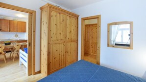 A room with a wooden wardrobe, a small window with a mirror, and a kitchen area visible through an open door.