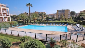 Ein rechteckiger Swimmingpool mit Fliesen umrandet, umgeben von einem Metallzaun. Entlang des Poolrands reihen sich Liegestühle.