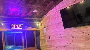Ett rum med ett träpanelat tak, en takfläkt, en stor TV som är monterad på väggen, en blå soffa, trästolar och ett fönster med en skylt som visar "ÖPPET".
