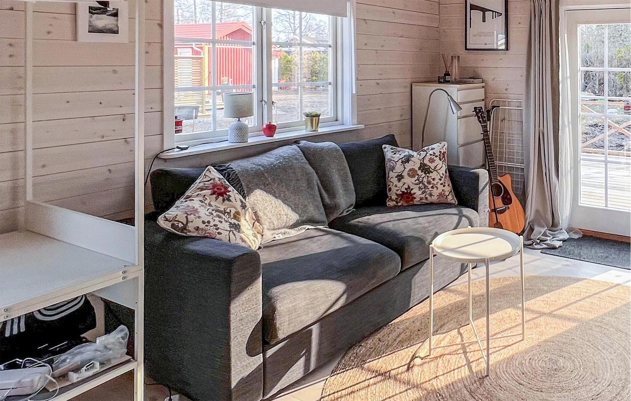 Una sala de estar acogedora con un sofá gris oscuro, cojines estampados, una mesita auxiliar blanca, una guitarra y una ventana con vistas a un edificio rojo y árboles.